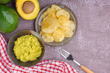 Organic avocado with seed, avocado halves, and fresh guacamole in a bowl with potato chips on a plate with cloth and cutlery on gray stone background. Top view. Concept of healthy fruit