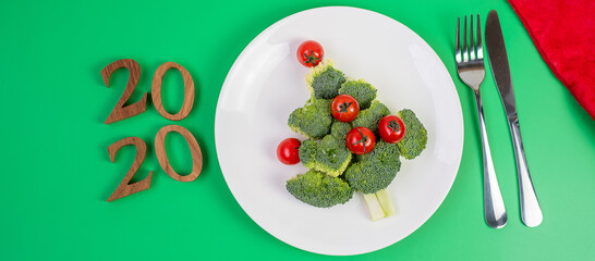 2020 Christmas tree of vegetables on white plate; Broccolis and tomatoes with fork and knife on green background. Merry Xmas and happy New Year party, New You, Healthy food and vegetarian concept