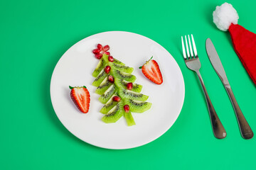 Christmas tree of vegetable and fruit; Kiwi, Strawberry and Pomegranate seed with fork and knife on green background. Merry Xmas and happy New Year party, New You, Healthy food and vegetarian