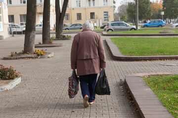 An old lonely elderly poor gray-haired woman with bags walks from the store or down the street. Rear view, face not visible.