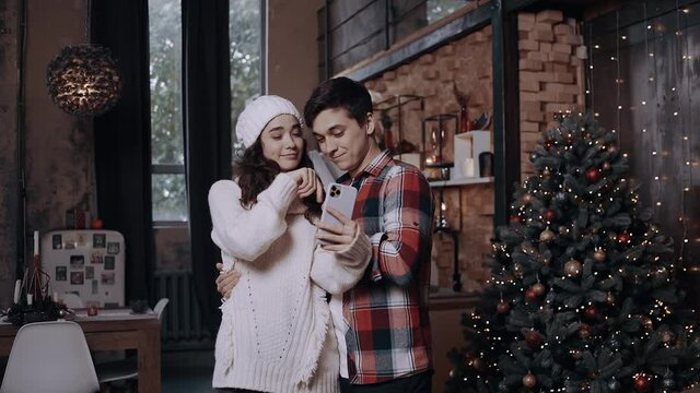 Young couple dressed in Christmas clothes, having fun and taking pictures with their phone near the Christmas tree. Happy winter holidays and hobby concept