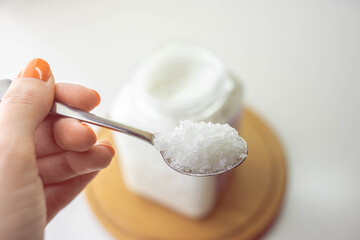 Female hand holds a spoon with white sea salt. Blurred background of jar and wooden board on white table