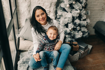 Happy mother with son sit on windowsill near Christmas tree and gift box.