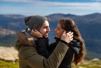 Young couple in love and in winter clothes hug and smile in front of the mountain range in a sunny day