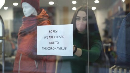 Young Caucasian woman in Covid face mask approaching to shop window and hanging announcement Sorry we are clothed due to the coronavirus. Trading closure due to infectious viral pandemic.