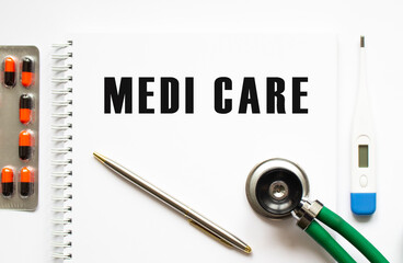 MEDI CARE is written in a notebook on a white table next to pills and a stethoscope.