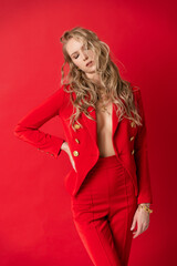 A beautiful blonde curly girl wearing a red pantsuit, unbuttoned on her braless big breasts sensually poses with her eyes closed on a red background. Advertising, fashionable, commercial design