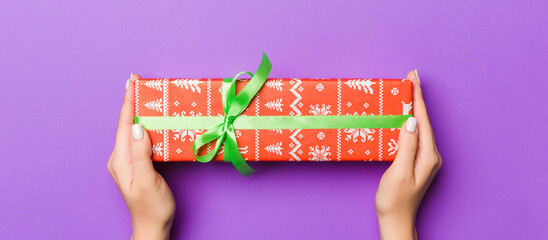 Flat lay of woman hands holding gift wrapped and decorated with bow on purple background with copy space. Christmas and holiday concept