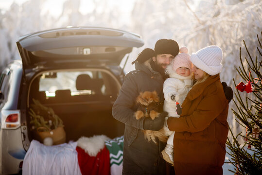 Young parents of the family are holding a little daughter and a dog on the background of Christmas decorations in the trunk of an SUV
