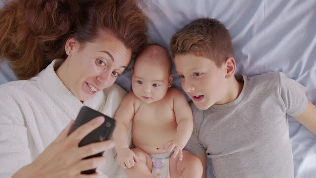 Happy mother with her kids are making a selfie or video call to father or relatives in a bed. Concept of technology, new generation,family, connection, parenthood, authenticity.