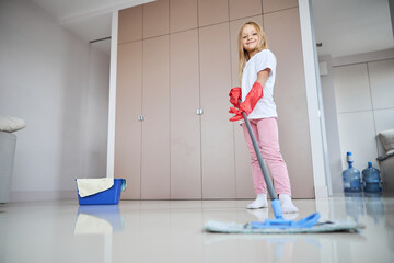 Charming small miss in casual clothes helping clean floor to her mom
