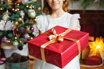 Selective focus. A girl in a white dress holds a red gift by the fireplace and Christmas tree.
