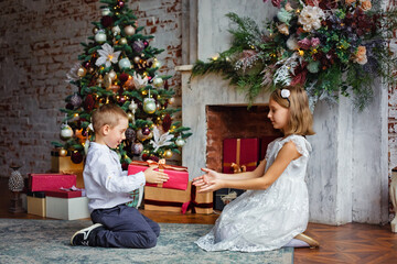 Obraz na płótnie Canvas Brother and sister are smartly dressed with a gift near the fireplace and Christmas tree with gifts. A boy gives a gift to a girl. Congratulations on the holiday.