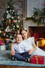 Fototapeta na wymiar Brother and sister smartly dressed with a gift near the fireplace and Christmas tree with gifts.