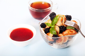 Minipancakes and blackberries in a glass cup, jam and a cup of tea on a white background.