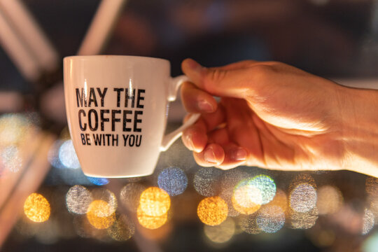 A hand holding a coffee cup is in focus with bokeh lights in the background, and has a funny quote that is a play on a famous sci-fi movie line. 