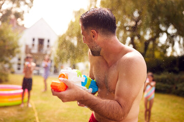 Family Wearing Swimming Costumes Having Water Fight With Water Pistols In Summer Garden