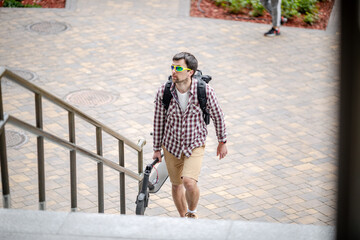 Young caucasian man in casual clothes with a backpack carries a folded electric scooter in his hand up the stairs of an office building. Man Carrying E-Scooter in folded position From Work