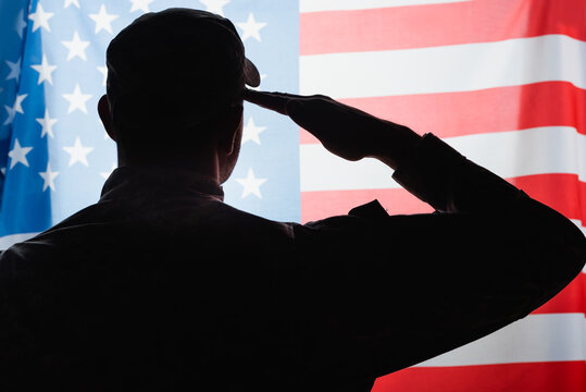 back view of patriotic military man in uniform and cap giving salute near american flag