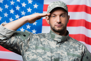 patriotic military man in uniform and cap giving salute near american flag on blurred background