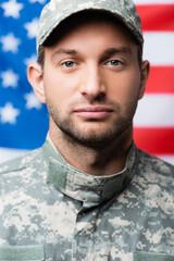military man in uniform and cap looking at camera near american flag on blurred background