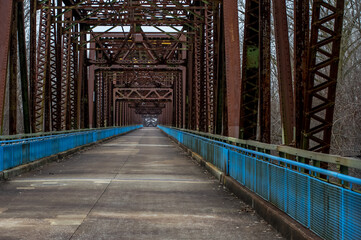 Old rusty steel truss bridge going into background with concrete deck and blue rails for pedestrians in winter