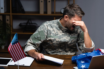 upset military man in uniform covering eyes and holding letter while screaming near gadgets and american flag