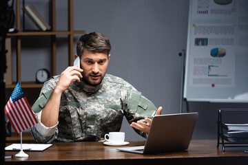 patriotic military man in uniform talking on smartphone near american flag, cup and laptop