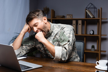 tired military man in uniform sitting with clenched fist while yawning near laptop