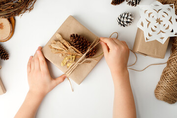 Child's hands making Christmas gift. Flat lay, top view, Kid hands wrapped gift box in craft paper, natural decoration details, zero waste packaging on white table. Eco-friendly concept
