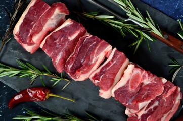 Raw meat ribs on a black stone plate, decorated with green sprigs of rosemary, Dark granite background. Raw Organic Beef Short Ribs