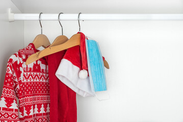 White and red christmas pajamas, hat, tights and face mask hanging on wooden hangers inside a white closet. Holidays during coronavirus pandemic..