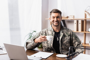 cheerful military man in uniform holding cup of coffee near laptop on desk