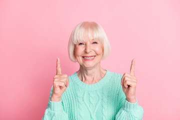 Photo portrait of pretty old lady pointing up at copyspace smiling wearing teal knitted sweater isolated on pastel pink color background