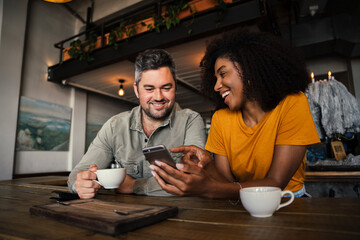 Happy couple laughing at images on social media holding smartphone drinking coffee in funky cafe 
