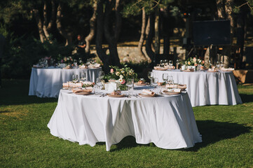 Large round tables with a white tablecloth and empty dishes, a bouquet of flowers stand on green...