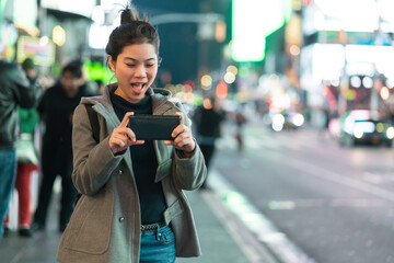 Fototapeta premium asian female traveller hand use smartphone and camera to capture photo in journey vacation night time nightlife in down town city street intersection light bokeh and crowd people tourists background