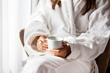 Girl holding a white coffee mug. In a white coat. Coffee in bed. Morning begins with coffee. Coffee in the hands of the bride