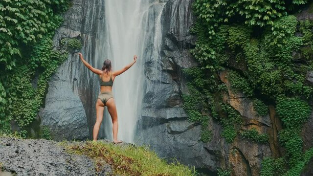 Young adventurous woman, successful and independent, confident and empowered female stands on top of rock and looks on waterfall with her hands raised. Beautiful accomplishment outdoors lifestyle