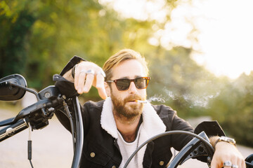 Fototapeta premium Portrait of a man smoking with rude expression while leaning against a handlebar of a motorcycle