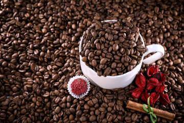 White cup full of coffee beans on a coffee beans background with red chocolate candy and red flowers. Morning espresso. Coffee mug. 