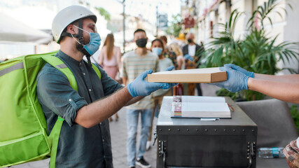 Delivery man wearing mask and protective gloves receiving pizza from hands of shop assistant while collecting his order from the pickup point during coronavirus lockdown