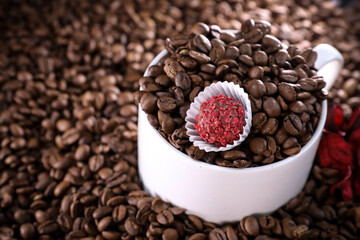 White cup full of coffee beans on a coffee beans background with red chocolate candy and red flowers. Morning espresso. Coffee mug. 