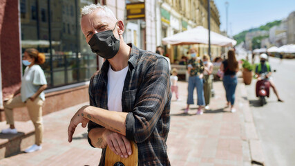 Portrait of middle aged man wearing mask holding a longboard and looking at camera. People collecting their orders from the pickup point during coronavirus lockdown in the background