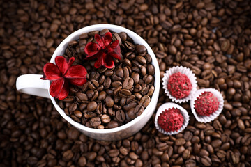 White cup full of coffee beans on a coffee beans background with red chocolate candy and red flowers. Morning espresso. Coffee mug. 