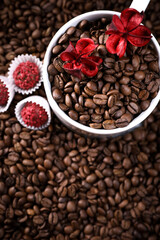 White cup full of coffee beans on a coffee beans background with red chocolate candy and red flowers. Morning espresso. Coffee mug. 