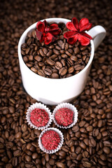 White cup full of coffee beans on a coffee beans background with red chocolate candy and red flowers. Morning espresso. Coffee mug. 