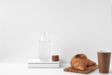 Fototapeta na wymiar Transparent bottle with cork stopper paper cup and books with fresh croissants on a white background. Natural and eco-friendly materials in interior decor. Copy space, mock up.