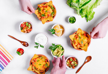 Nachos with chicken, mushroom and melted cheese in portion bowls served with assorted toppings on white table. Salsa, avocado, sour cream, lemons, green onion. Top view. Mexican food
