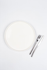 white round ceramic plate with a knife and fork on a white background. Look from above